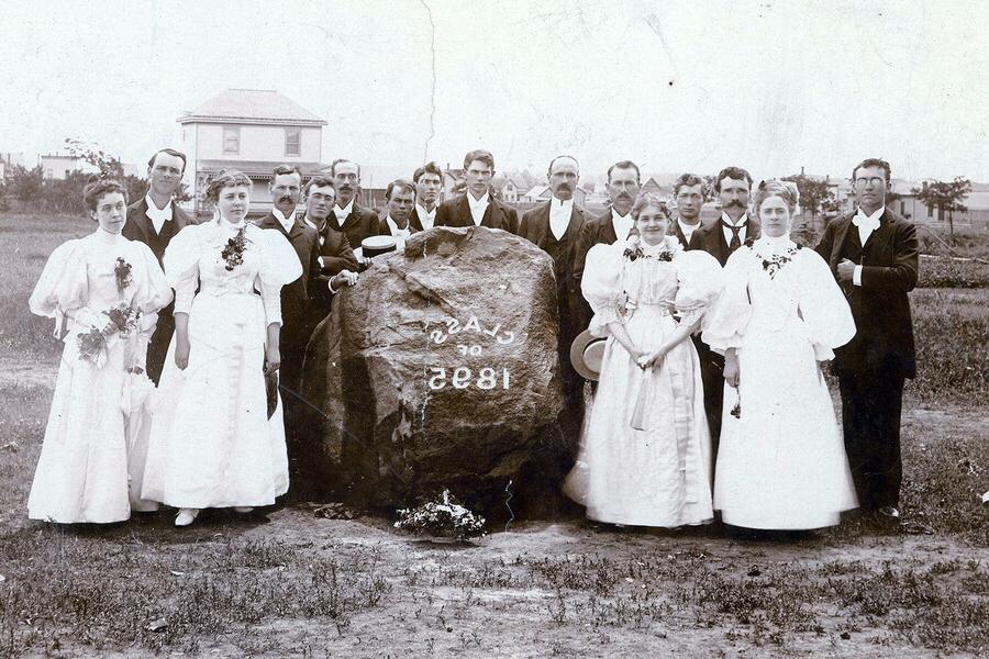 Class of 1895 included four women standing around a large rock with male students standing behind the rock. 